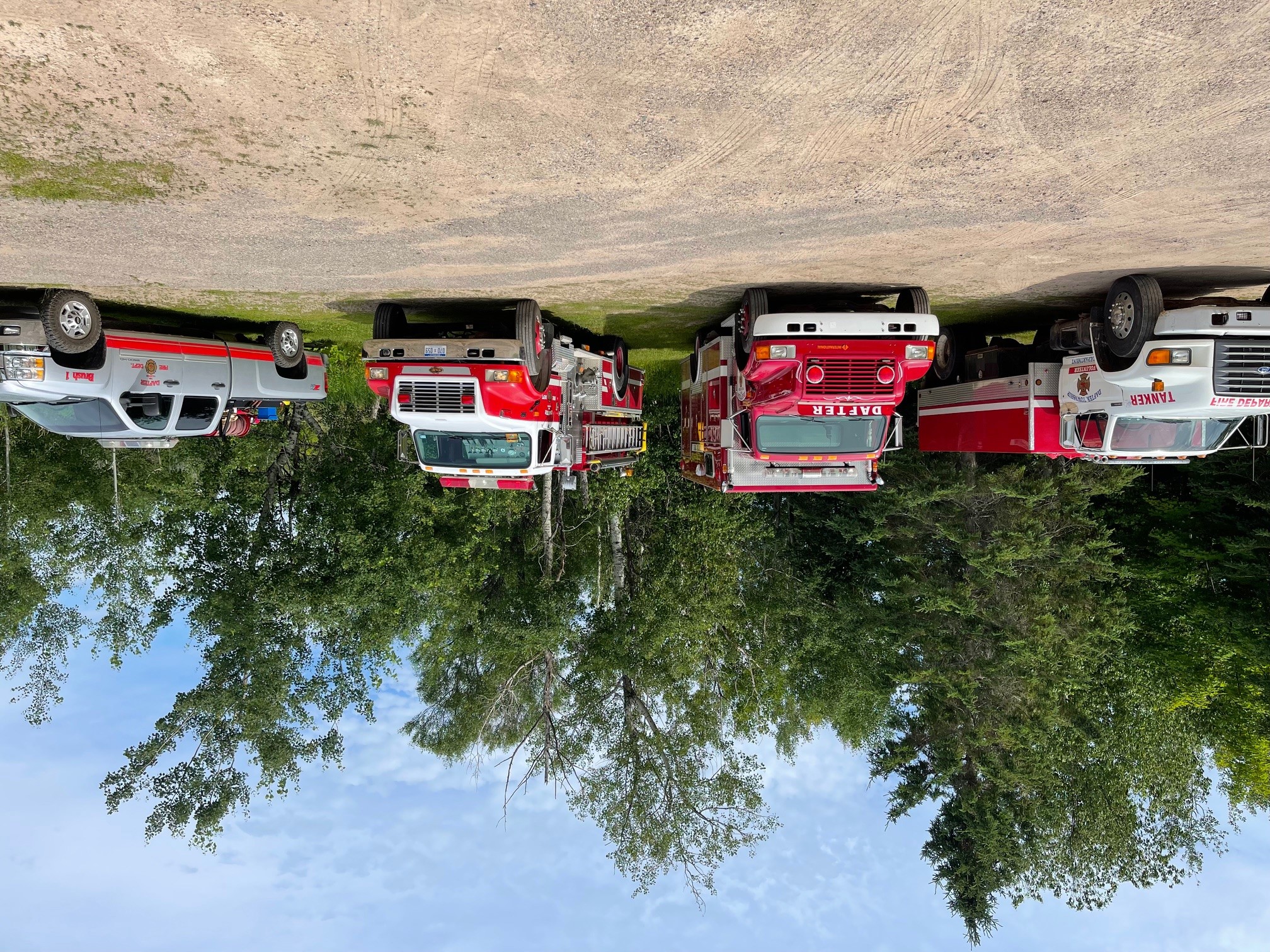 Row of fire department trucks parked in front of a stand of trees.