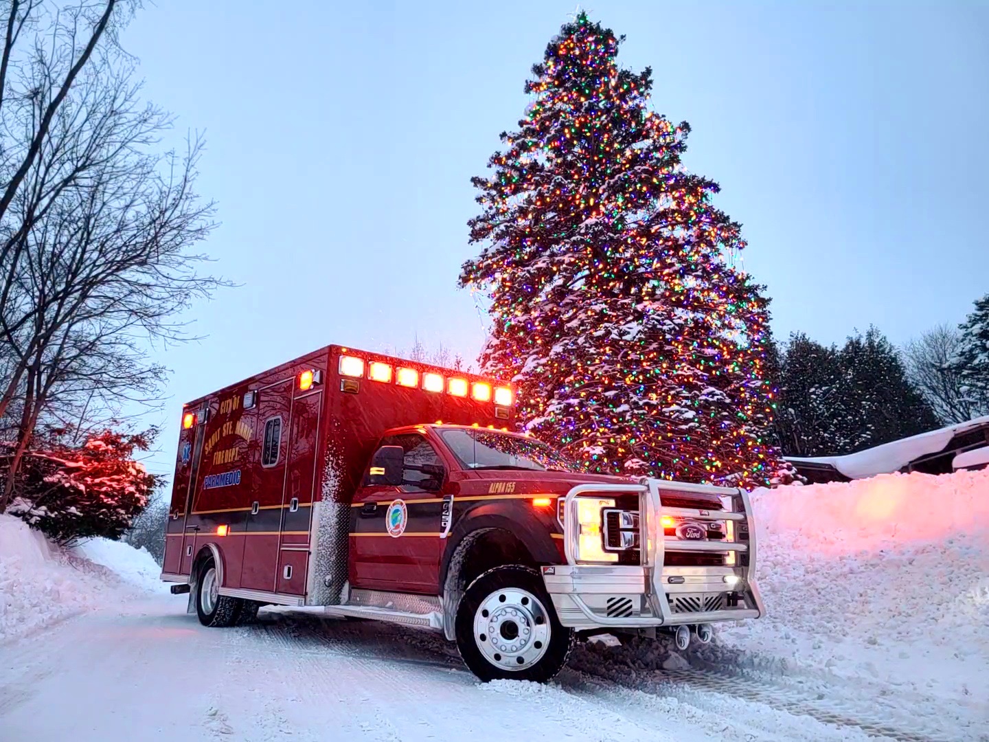 Ambulance with its emergency lights on, parked in the snow beside a lighted Christmas tree.