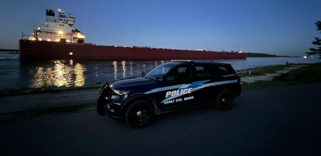 Sault Ste. Marie police SUV at dusk beside the St. Marys River, with a freighter behind it.