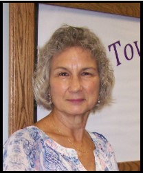Woman with gray curly hair wearing a light blue patterned top, standing in front a partially visible sign.