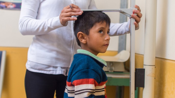 Healthcare provider measuring a young boy's height using a wall-mounted measuring scale during a medical examination.