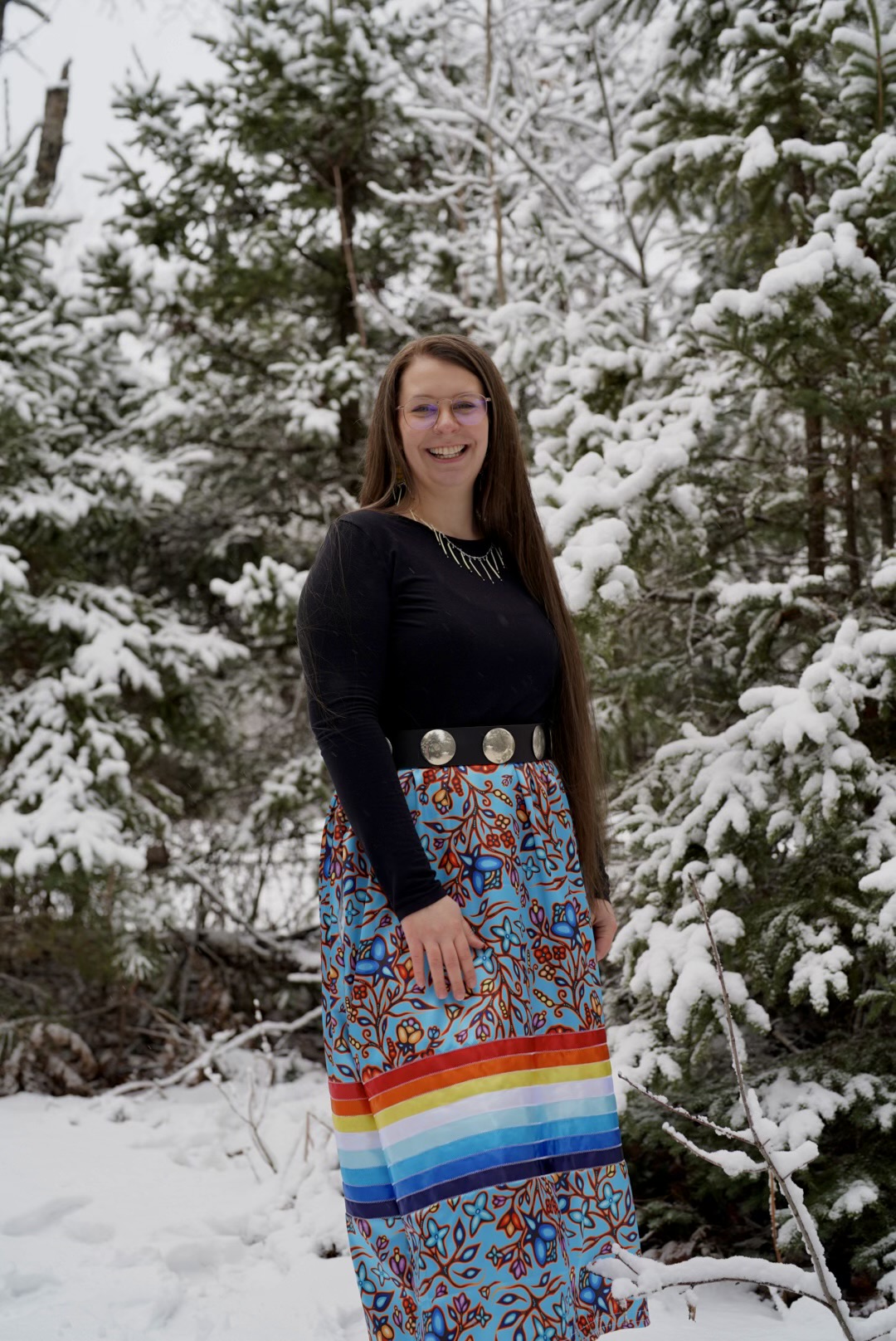 Young woman with long brown hair and glasses smiling at camera in snowy forest surrounded by snow-covered evergreen trees.