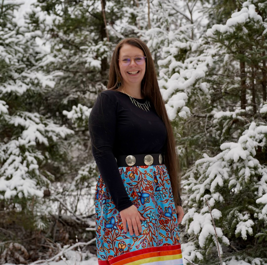 Woman with long brown hair and glasses smiling in front of snow-covered evergreen trees.