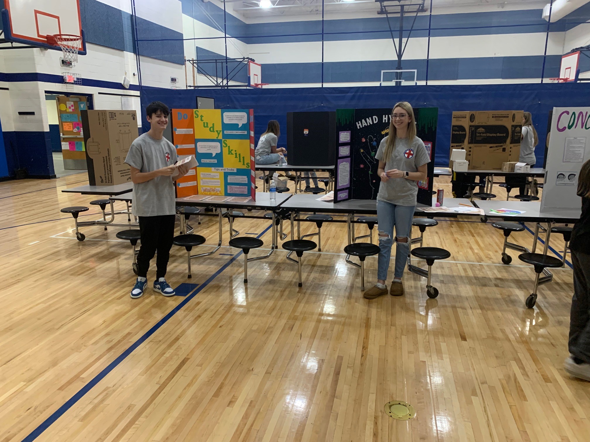Two students present science projects at tables in a school gymnasium with blue and white striped walls and basketball hoops.