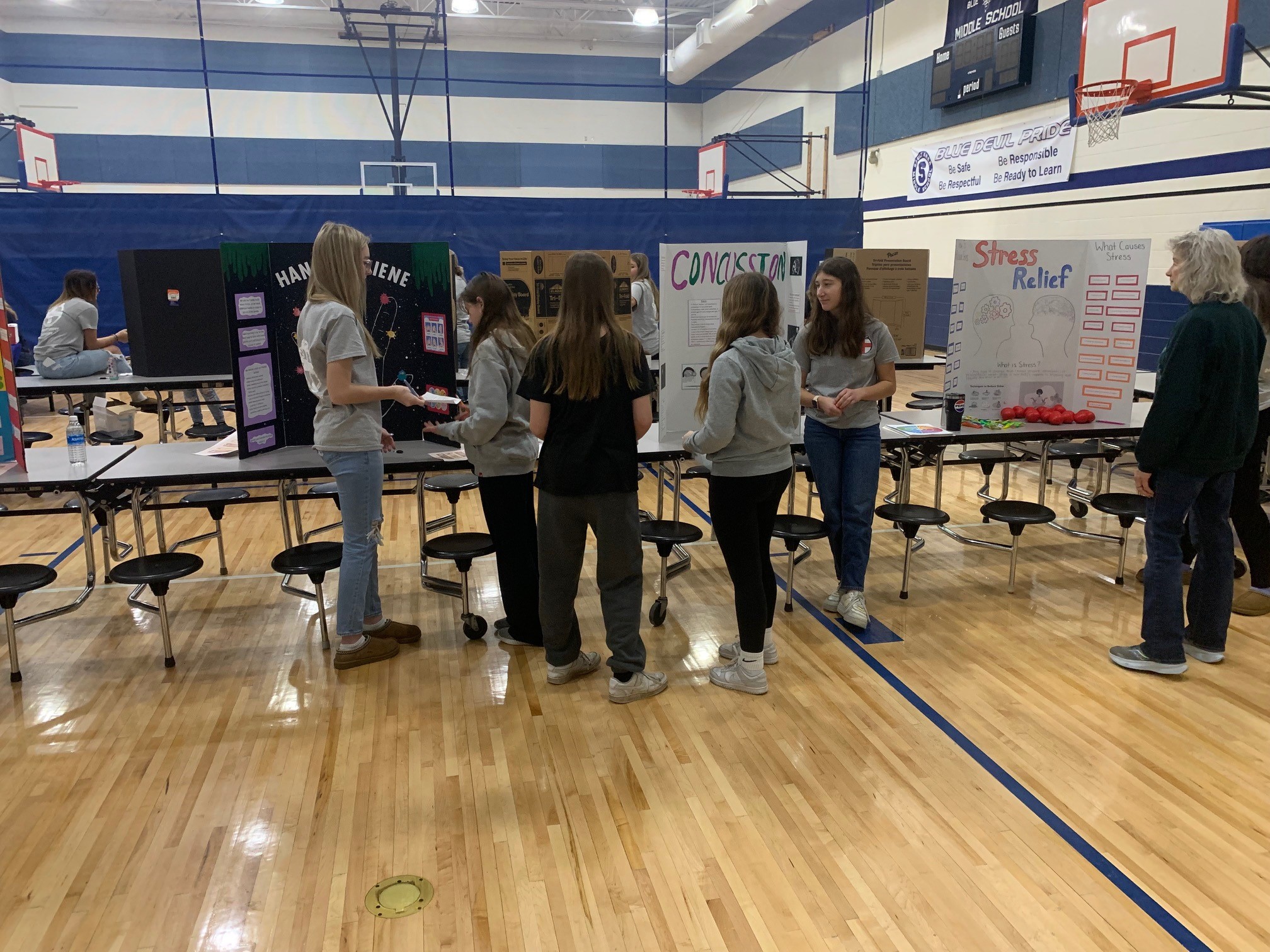 Middle school students present science fair projects on display boards in a gymnasium while visitors examine their work.