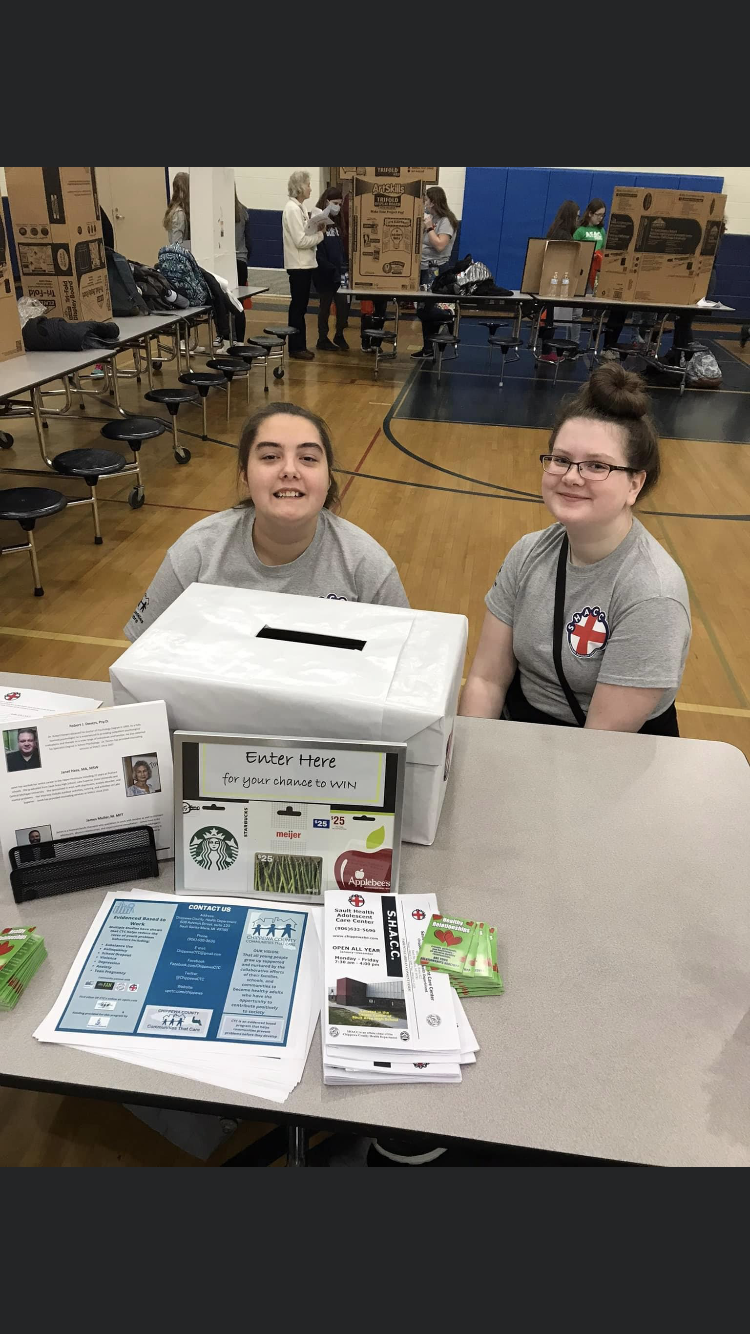 Two young women at a table with a raffle entry box and informational materials at a community event in a gymnasium.