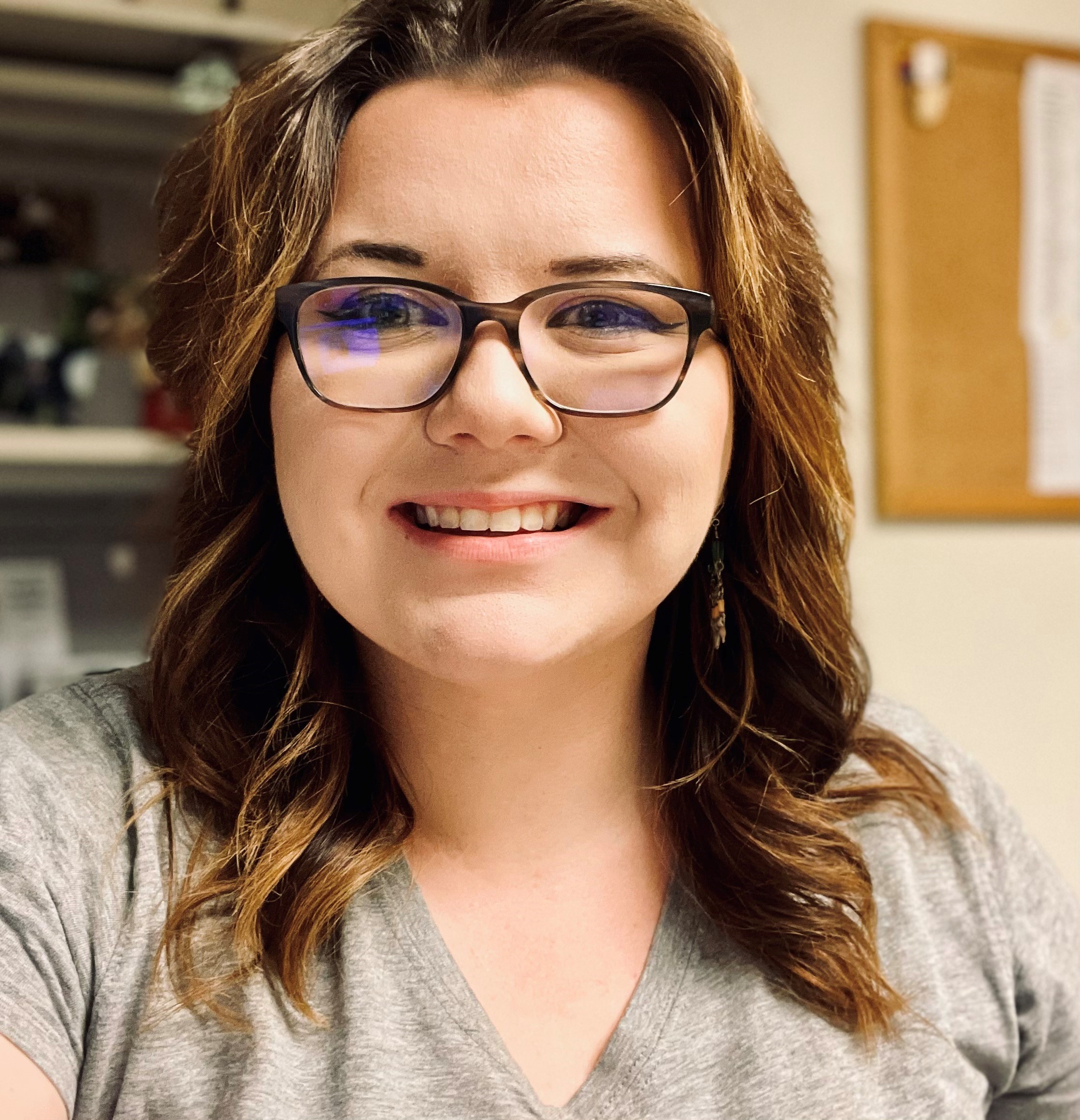 Woman with shoulder-length reddish-brown hair and black-framed glasses smiling at camera in an office setting