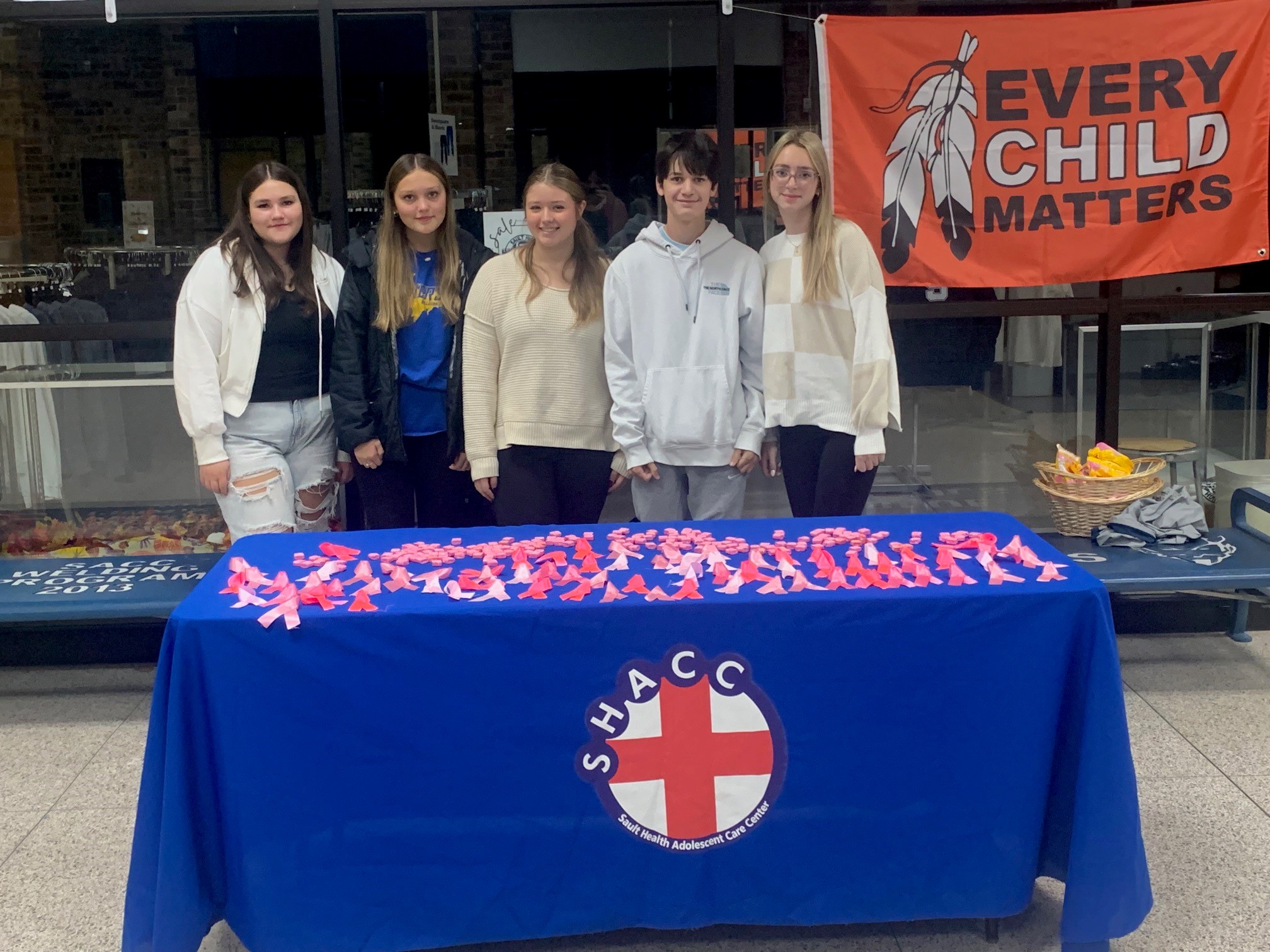 Five young people standing behind a blue table with red and white paper cutouts and an 'Every Child Matters' banner in the background.