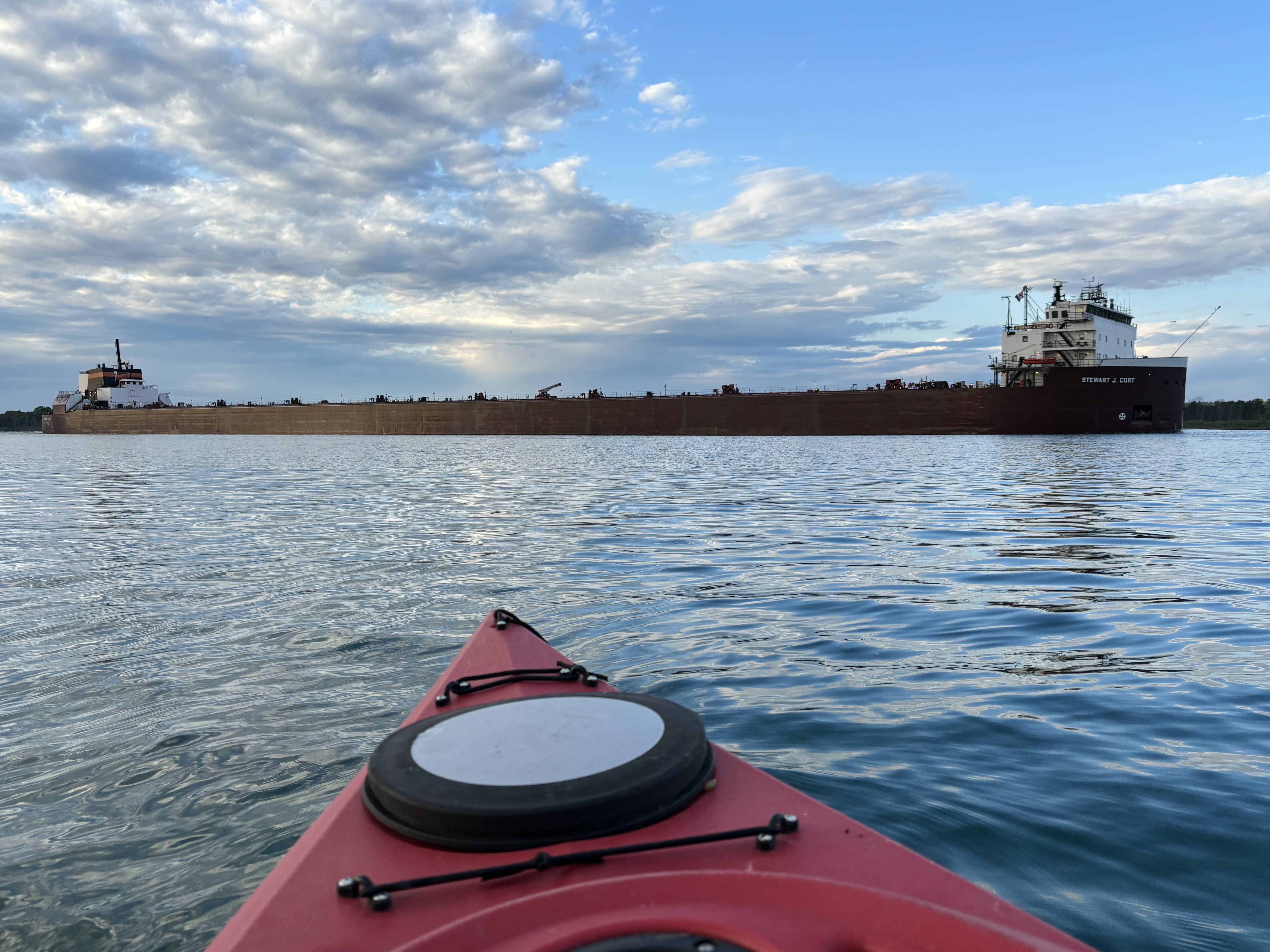 Red kayak in foreground on river with large cargo ship passing in the distance under cloudy sky.