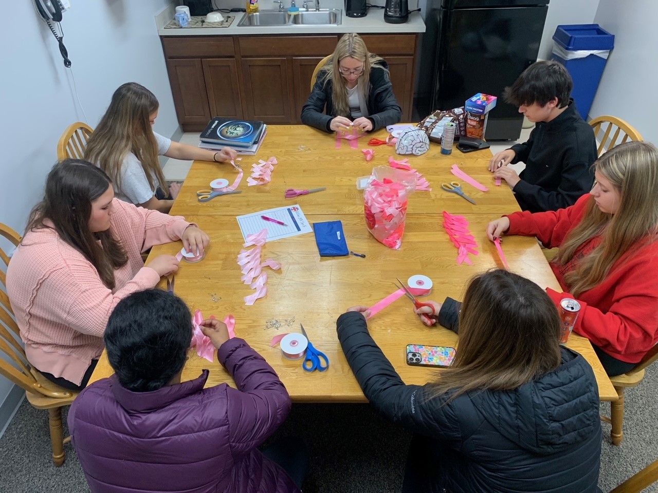 An adult supervisor and six young people sit around a wooden table creating pink paper flower decorations with scissors and ribbon.