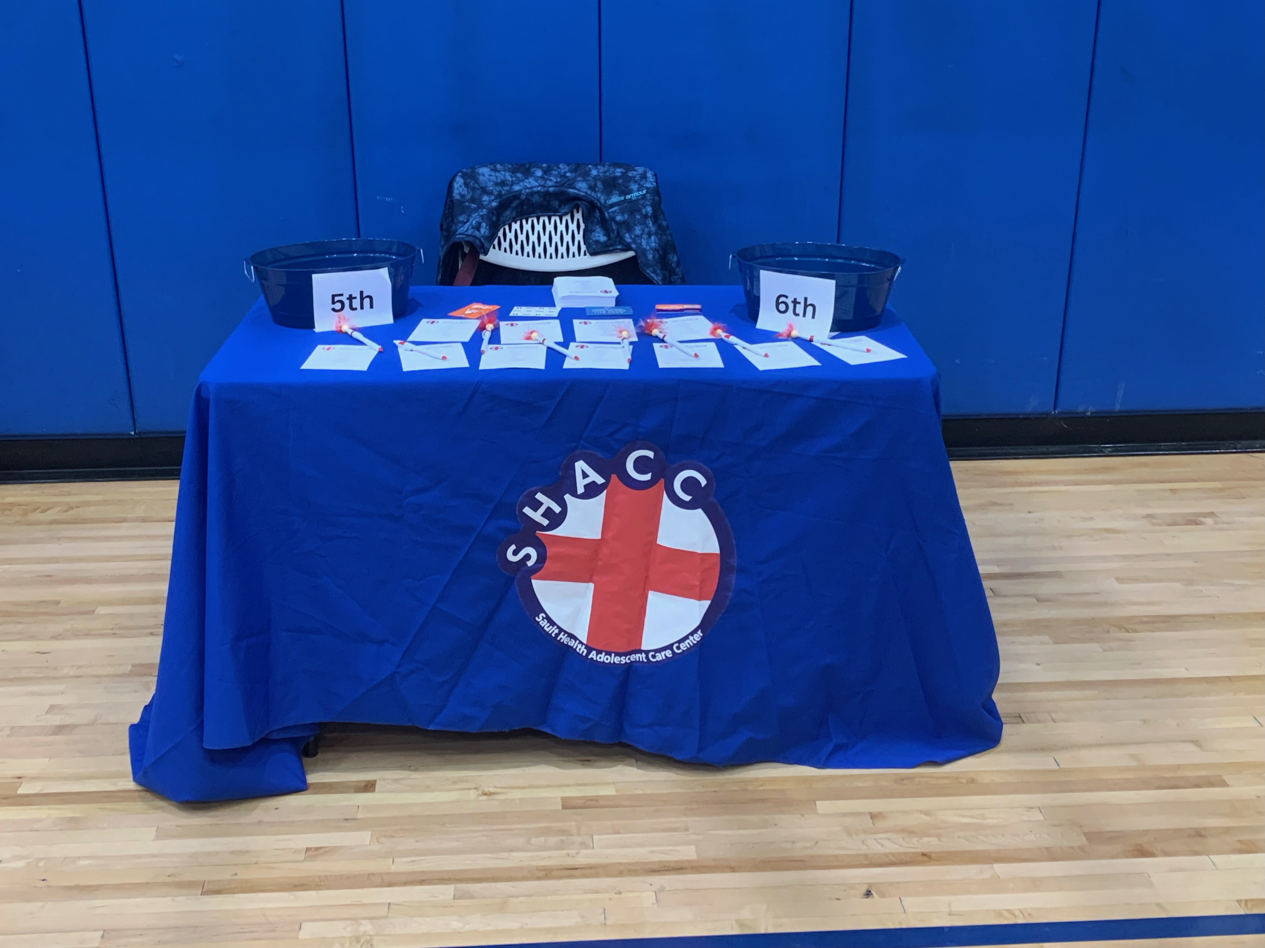 Blue table with SHACC logo displays educational materials organized by grade level 5th and 6th grade sections.