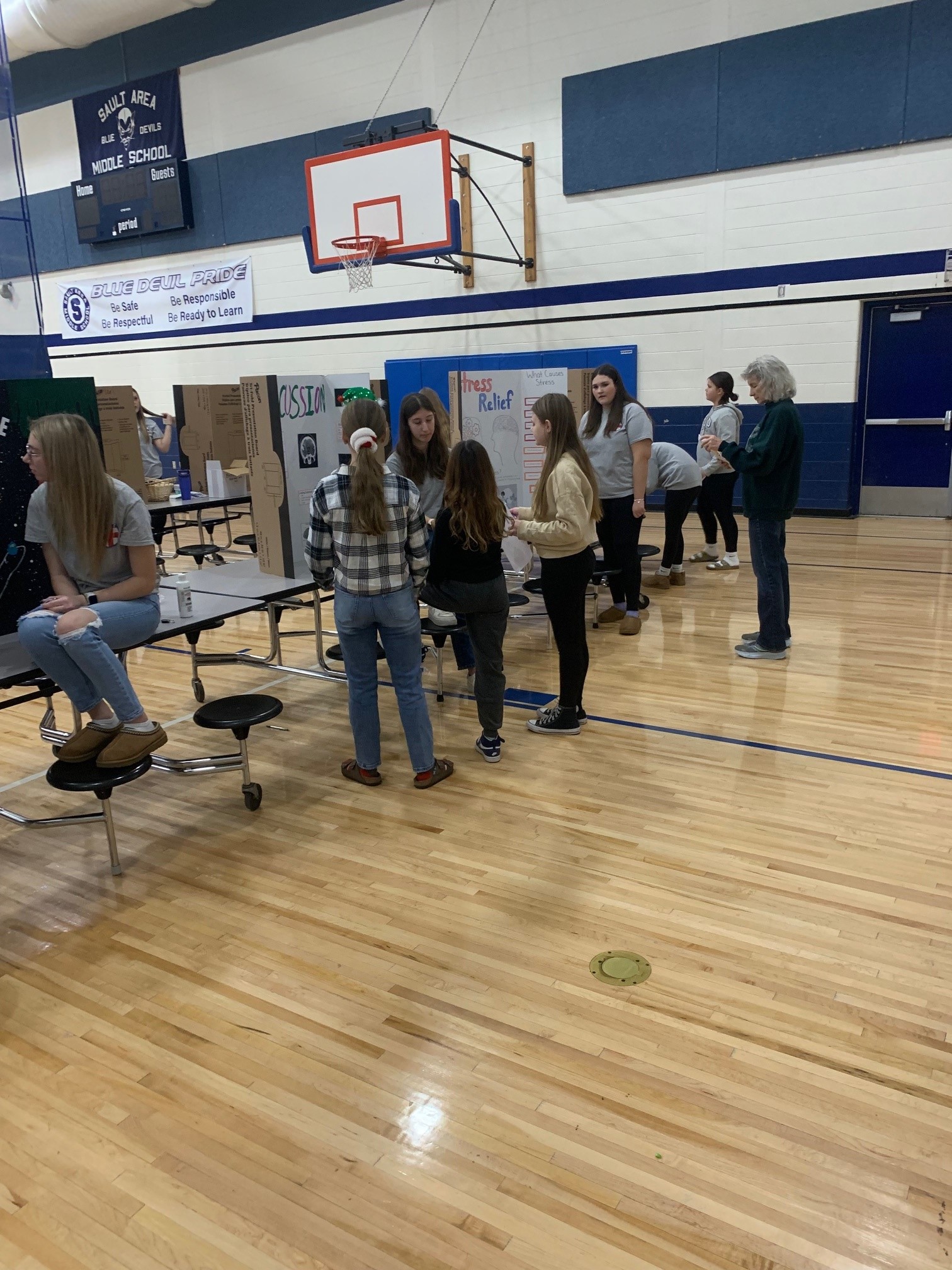 Middle school students gathered in a gymnasium viewing informational posters about community service projects displayed on cardboard boards.